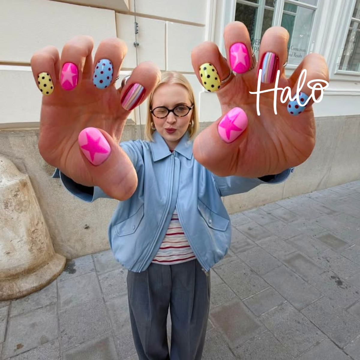Colourful nail art featuring bright pink stars, yellow and blue polka dots and multicoloured stripes, displayed by a woman posing outdoors.