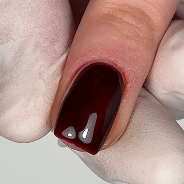 A close-up of a fingernail painted with a glossy deep burgundy nail polish, held by a hand wearing a white glove.
