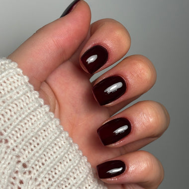 A close-up of a hand with manicured nails painted in a glossy deep burgundy shade, wearing a cream-colored knit sweater against a neutral gray background.