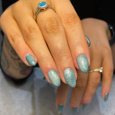 Close-up of a hand with blue glittery nail polish and a ring on a neutral background