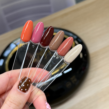A hand holds a fan of six nail color swatches on clear plastic sticks, showing shades of orange, pink, burgundy, taupe, dusty rose, and cream, with a black nail lamp in the background.