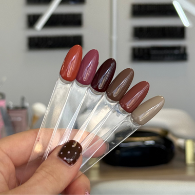 A hand holds a fan of six clear nail polish sample sticks in different muted shades, including terracotta, burgundy, plum, taupe, brown, and beige, with a blurred nail salon background.
