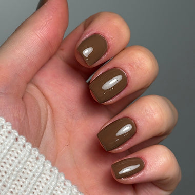 A close-up of a hand with a glossy medium brown nail polish manicure, wearing a white knit sweater against a neutral gray background.