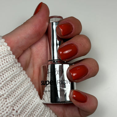 A hand with glossy burnt orange-red nail polish holding a bottle of Huda Super Rich Gel Nail Polish in the same shade, against a plain light background.