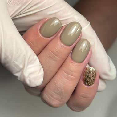 Close-up of manicured fingernails with a glossy taupe-beige polish on three nails and one glittery gold accent nail, shown with a white gloved hand holding the fingers.