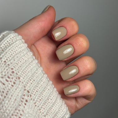 A close-up of a hand with glossy, short oval nails painted in a warm taupe-beige shade, wearing a white knit sweater against a plain gray background.