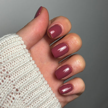 A close-up of a hand with glossy mauve-brown manicured nails, wearing a white knit sweater cuff against a plain gray background.