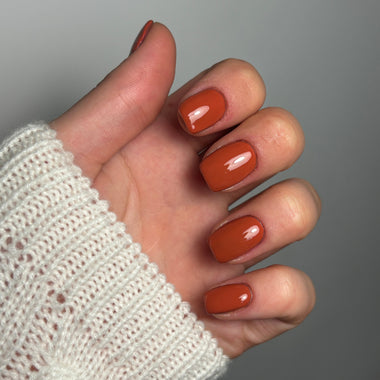 A close-up of a hand with glossy warm rust-orange manicured nails, wearing a cream knit sleeve against a plain gray background.