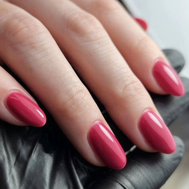 A close-up of a hand with long, almond-shaped nails painted in a glossy, solid berry pink shade, resting on a black surface.