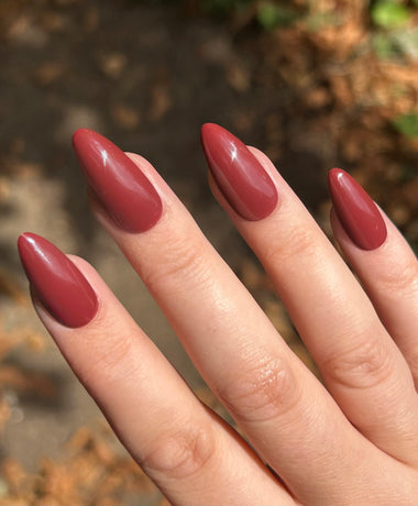 Close-up of a hand with almond-shaped nails painted in a glossy red-brown color against a blurred natural background.