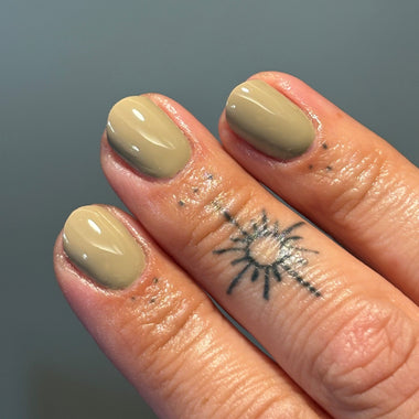 Close-up of a hand with manicured nails painted in a warm beige/taupe color, showing a small black sun tattoo on the ring finger against a soft gray-blue background.