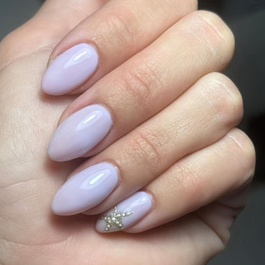 Hand with lavender-colored nails, one of which has gold glitter, against a neutral background.