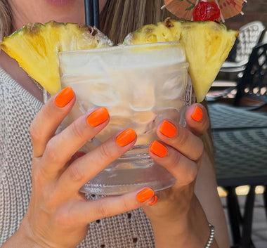 Person holding a cocktail with pineapple slices, wearing bright orange nail polish.