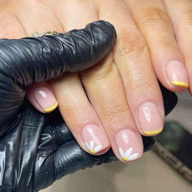 Nails with white floral designs and yellow accents, being held by a person wearing black gloves.