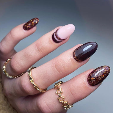 Hand with dark brown and pink nail polish wearing gold rings on a blurred background