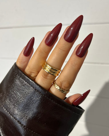 Hand with maroon nail polish wearing a black glove and gold rings on a white background