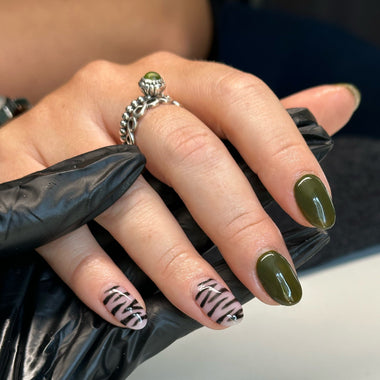 Hand with green nail polish and zebra pattern, wearing a silver ring, against a blurred background.