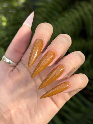 Hand with long, almond-shaped, brown acrylic nails against a blurred green background