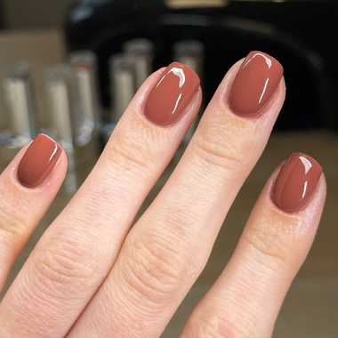 A close-up of a hand with fingernails painted in a glossy dusty rose or mauve shade, with blurred nail polish bottles in the background.