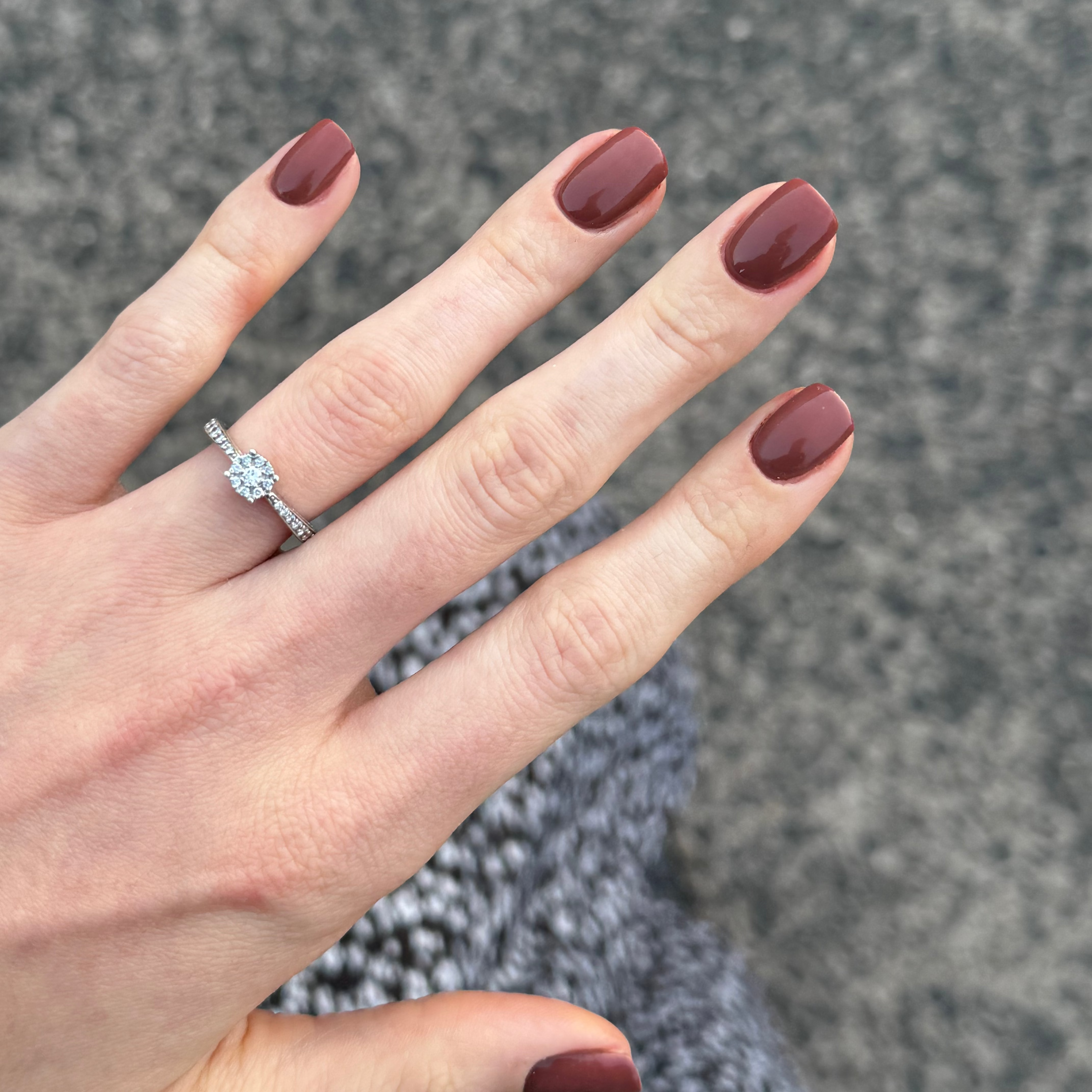 A close-up of a hand with dark burgundy  brown nail polish wearing a silver ring with a round clear gemstone, set against a blurred gray textured background.