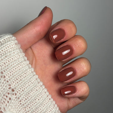 A close-up of a hand with glossy brown manicured nails, wearing a white knit sweater cuff, set against a solid gray background.