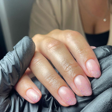 Hand with pink glittery nails held by a person wearing black gloves.
