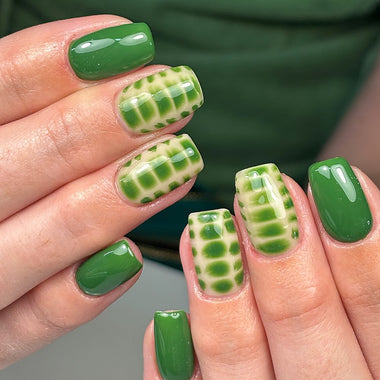 Close-up of hands with green nail polish, some checkered pattern, against a blurred green background.