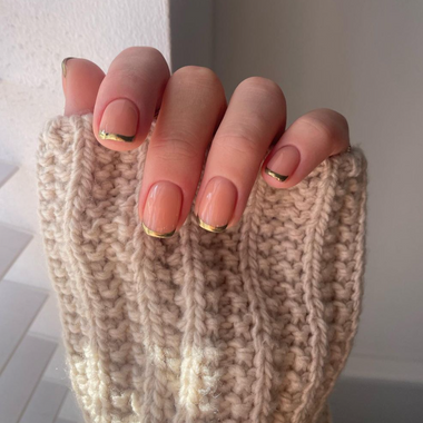 Hand wearing a beige knitted sleeve with gold nail polish on a neutral background