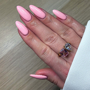 Hand with pink nails and a ring featuring a large diamond and pink gemstone on a light wooden surface.